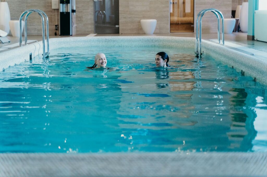 Two people are swimming and talking in an indoor pool with modern decor and metal handrails.