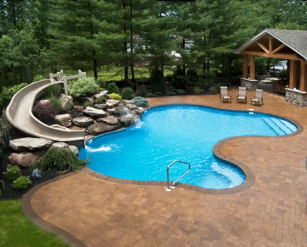 Backyard swimming pool with a curved water slide, brick patio, lounge chairs, and a covered seating area, surrounded by trees and landscaping.