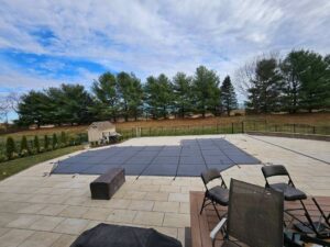 Rectangular pool covered with a safety cover in a backyard patio, surrounded by trees and outdoor furniture under a partly cloudy sky.